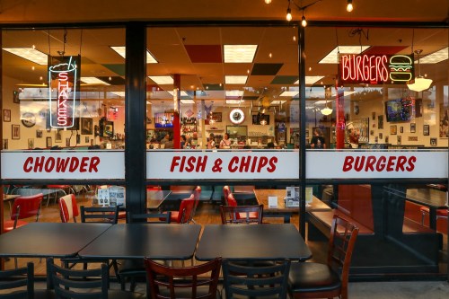 A restaurant window displaying signs for chowder, fish & chips, and burgers, with a cozy interior and seating.