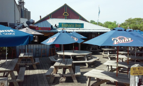 Outdoor seating area with wooden picnic tables and blue umbrellas in front of a restaurant named Barnacle Bud's.