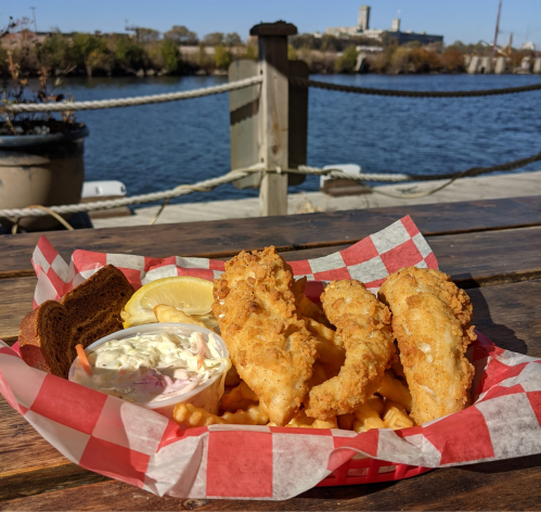 A basket of fried fish fillets with fries, coleslaw, lemon, and a slice of bread, set by a waterfront view.
