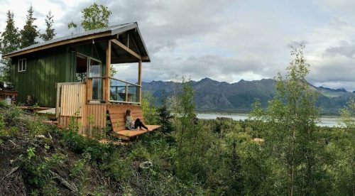 A green cabin overlooks a lush landscape with mountains and a lake under a cloudy sky.