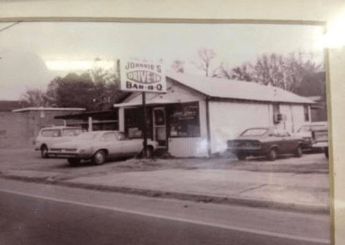 Black and white photo of Johnny's Drive-In Bar-B-Q, featuring vintage cars parked outside a small, rustic building.