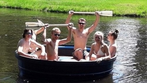 A group of five friends in a small boat on a river, smiling and holding paddles, enjoying a sunny day.