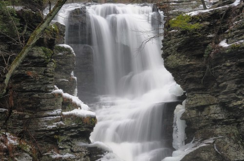 A serene waterfall cascading over rocky cliffs, surrounded by snow and bare trees in a tranquil natural setting.
