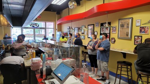 A busy restaurant interior with customers in line, staff preparing food, and colorful decor on the walls.