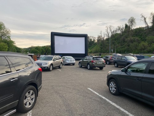 An outdoor drive-in movie setup with an inflatable screen and parked cars in a lot surrounded by greenery.