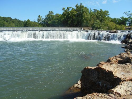A serene waterfall cascades over a dam, surrounded by lush greenery and rocky shoreline under a clear blue sky.