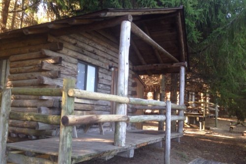 A rustic wooden cabin with a porch, surrounded by trees in a natural setting.