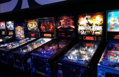 A row of colorful pinball machines, including "Jurassic Park," in a dimly lit arcade setting.