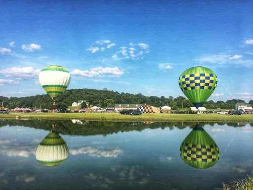 Two colorful hot air balloons reflect in a calm pond under a clear blue sky, surrounded by green hills and tents.