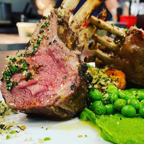 A close-up of a perfectly cooked lamb chop, garnished with herbs, alongside green peas and a vibrant green puree.