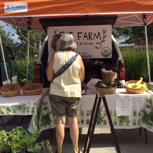 A woman browses fresh produce at a local farm stand under an orange canopy, with baskets of vegetables displayed.