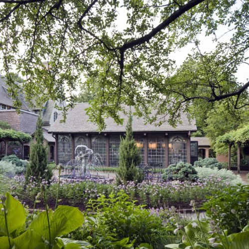 A serene garden with a fountain, surrounded by lush greenery and flowering plants, framed by trees and a cozy building.