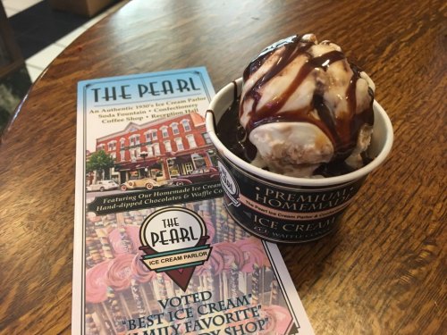 A bowl of ice cream topped with chocolate sauce, next to a menu for The Pearl Ice Cream Parlor on a wooden table.