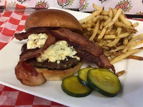 A juicy burger topped with bacon and blue cheese, served with crispy fries and pickles on a white plate.