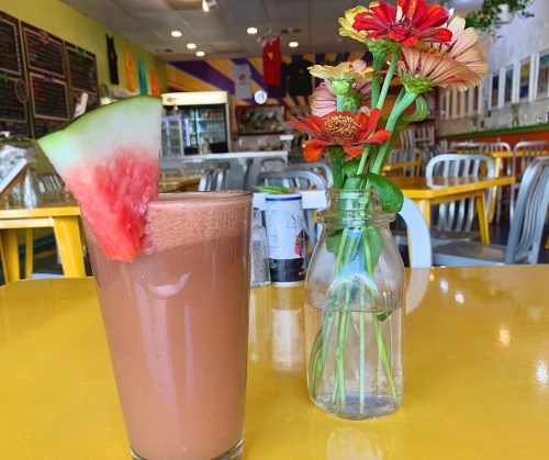 A chocolate smoothie with a watermelon slice beside a vase of colorful flowers on a bright yellow table.