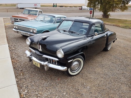 A classic black car parked in front of two vintage cars, set against a gravel lot and open landscape.