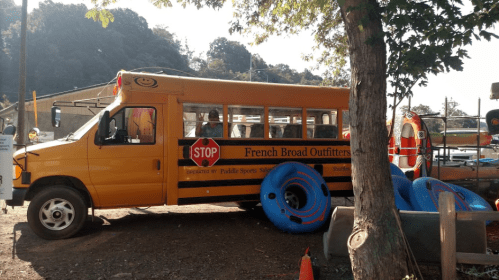 A yellow bus labeled "French Broad Outfitters" parked near inflatable tubes and trees on a sunny day.