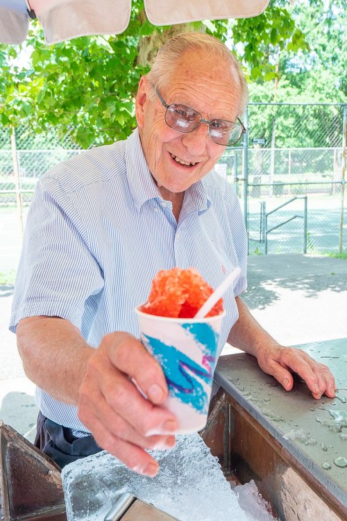 An elderly man smiles while holding a colorful snow cone at a outdoor stand.