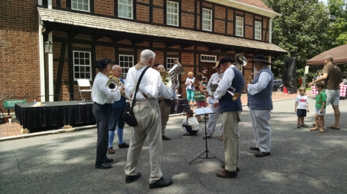 A group of musicians in period costumes play brass instruments outdoors near a historic building.