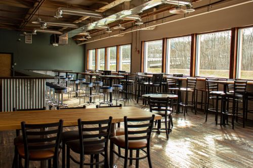 A spacious, modern interior with wooden floors, tables, and stools, featuring large windows and natural light.
