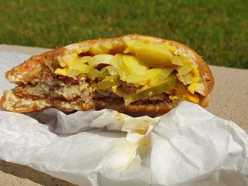 A partially eaten sandwich with meat, yellow mustard, pickles, and onions, resting on a white wrapper outdoors.
