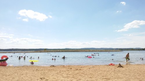 A sandy beach with people swimming and playing in the water under a clear blue sky with a few clouds.