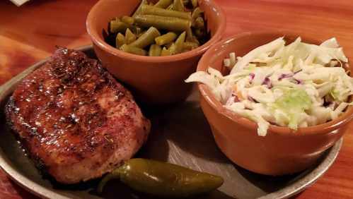A plate with grilled pork, green beans, coleslaw, and a jalapeño pepper in a rustic setting.
