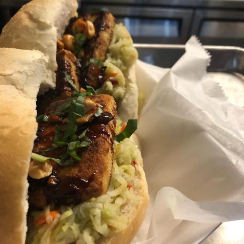 A close-up of a sandwich filled with grilled chicken, slaw, and garnished with herbs and peanuts, served in a bun.