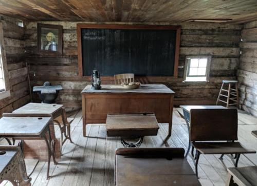 A rustic classroom with wooden walls, a chalkboard, a teacher's desk, and old-fashioned student desks and chairs.