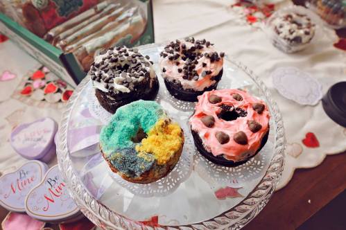 A glass cake stand with four colorful frosted cupcakes topped with chocolate chips and sprinkles.