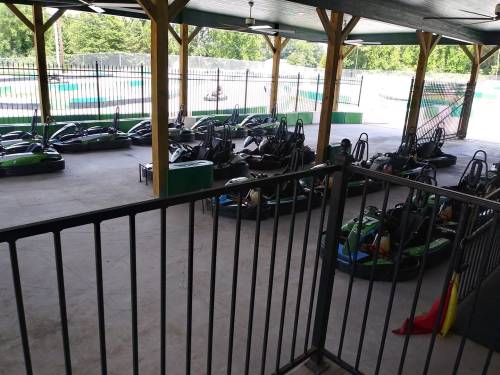 A view of go-karts lined up under a covered area at a racetrack, with a fence and trees in the background.
