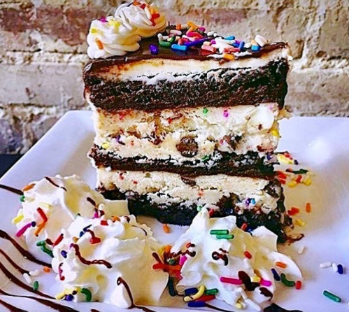 A tall slice of layered cake with chocolate, vanilla, and colorful sprinkles, topped with whipped cream and chocolate drizzle.
