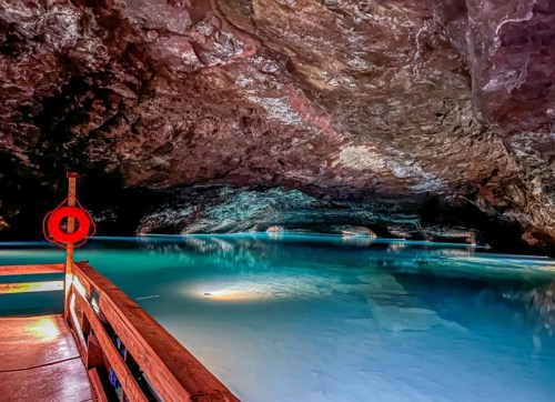 A serene underground cave with a calm blue pool of water and a wooden walkway, illuminated by soft lights.