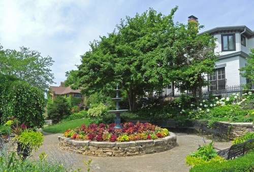 A landscaped garden featuring a fountain surrounded by colorful flowers and greenery, with a house in the background.