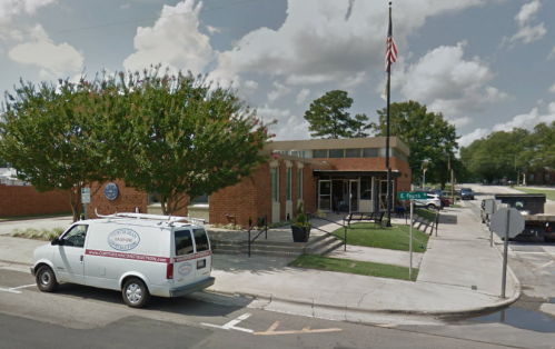 A brick building with a flagpole, surrounded by trees, and a van parked nearby on a sunny day.