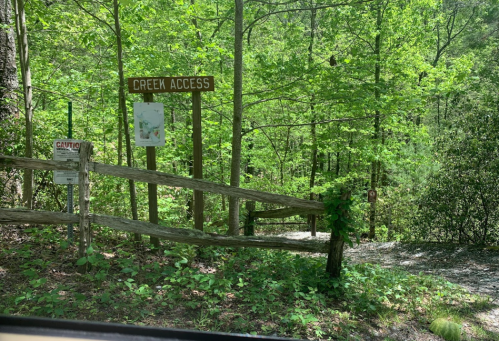 A wooden sign reading "Creek Access" near a forested area with greenery and a gravel path.
