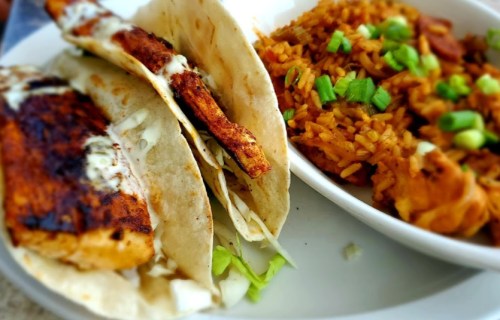 Three grilled fish tacos with cabbage and sauce, served alongside a bowl of seasoned rice topped with green onions.