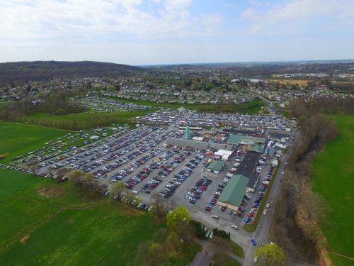 Aerial view of a large parking lot filled with cars, surrounded by green fields and residential areas in the background.