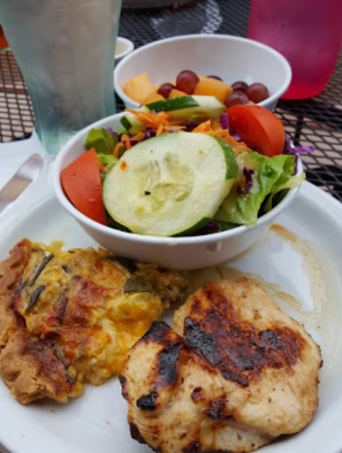 A plate with grilled chicken, a slice of frittata, and a fresh salad with cucumbers and mixed fruits on the side.
