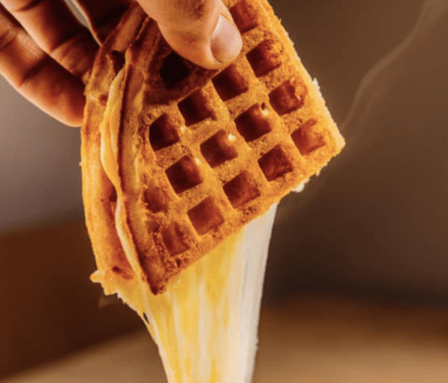 A hand holding a golden waffle with melted cheese stretching from it, against a warm background.