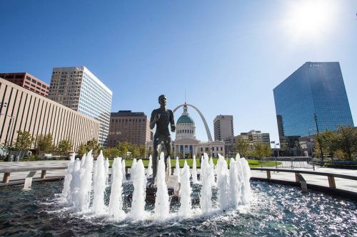 A fountain with a statue in the foreground, surrounded by buildings and a clear blue sky in a cityscape.