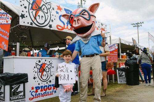A child in a pig costume holds a sign saying "EAT MOR PORK!" next to a person in a large pig mascot head at a fair.