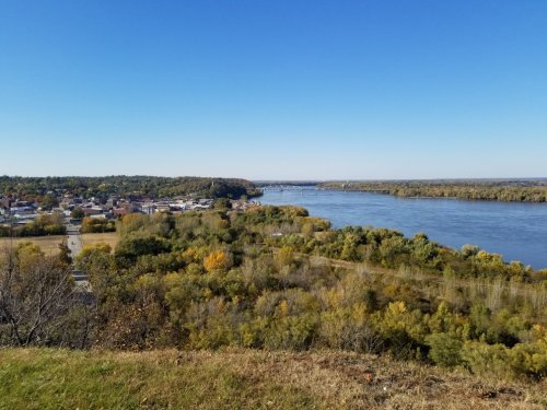 A scenic view of a river winding through a town, surrounded by trees and clear blue skies.