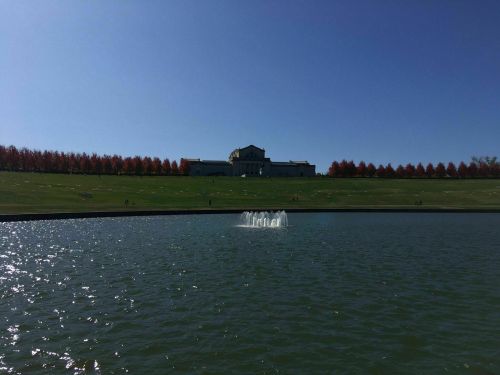 A serene lake with a fountain, surrounded by green grass and trees with autumn foliage, under a clear blue sky.