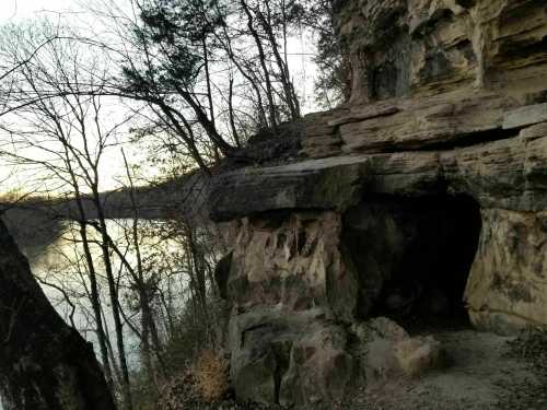 A rocky cliffside overlooking a calm river, surrounded by bare trees at sunset.