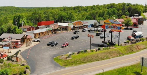 Aerial view of a colorful roadside attraction with shops, a fudge factory, and parked cars surrounded by greenery.