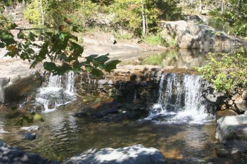 A serene waterfall cascades over rocks into a calm pool, surrounded by lush greenery and trees.