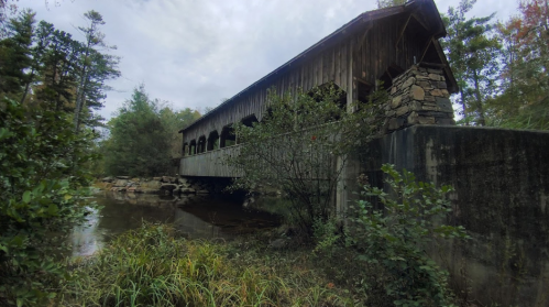 A rustic covered bridge spans a calm stream, surrounded by trees and greenery under a cloudy sky.