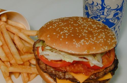 A sesame seed bun burger with lettuce, tomato, cheese, and a side of fries, next to a drink in a blue cup.