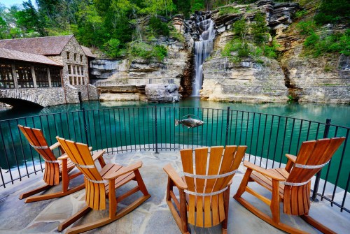 A serene outdoor scene featuring wooden chairs overlooking a tranquil lake and waterfall surrounded by lush greenery.